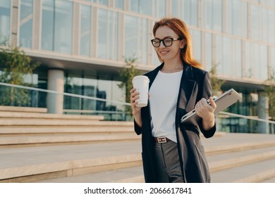 Beautiful Stylish Redhead Businesswoman With Laptop Drinking Coffee From Disposable Paper Cup While Going To Work In Morning, Female Business Professional Walking Outdoors On City Street And Smiling