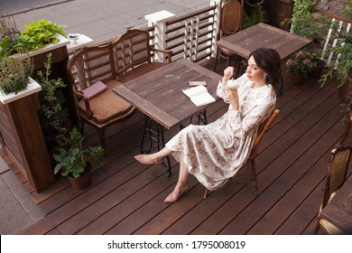 Beautiful Stylish Woman In Beige Dress Drinking Coffee Outdoors At The Cafe. Portrait Of Happy Female In Open Air Cafe Not  Looking At Camera 