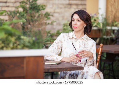 Beautiful Stylish Woman In Beige Dress Drinking Coffee Outdoors At The Cafe. Portrait Of Happy Female In Open Air Cafe Not  Looking At Camera 