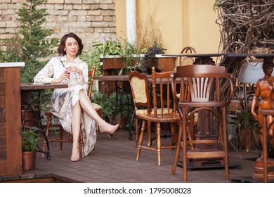 Beautiful Stylish Woman In Beige Dress Drinking Coffee Outdoors At The Cafe. Portrait Of Happy Female In Open Air Cafe Not  Looking At Camera 