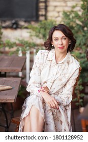 Beautiful Stylish Woman In Beige Dress Drinking Coffee Outdoors At The Cafe. Portrait Of Happy Female In Open Air Cafe Not  Looking At Camera 