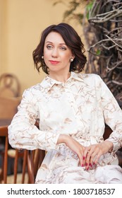 Beautiful Stylish Woman In Beige Dress Sitting At  Cafe Outdoor. Portrait Of Happy Female In Open Air Cafe, Not  Looking At Camera 