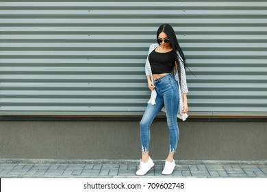 Beautiful stylish woman in a fashionable white cloak with a black T-shirt and blue jeans with a high waist with white shoes near the metal wall in the street