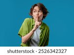 Beautiful stylish woman holding finger on lips, blue studio background. Pretty lady with gesture of shhh, secret, silence, conspiracy, gossip, rumors concept. High quality