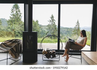 Beautiful stylish woman reading book on chair at fireplace with firewood on background of mountain hills. Young female in casual clothes relaxing in modern chalet with amazing view from window - Shutterstock ID 1950279142