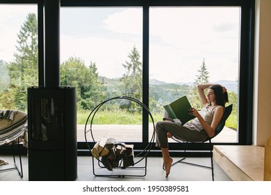 Beautiful stylish woman reading book on chair at fireplace with firewood on background of mountain hills. Young female in casual clothes relaxing in modern chalet with amazing view from window - Shutterstock ID 1950389581