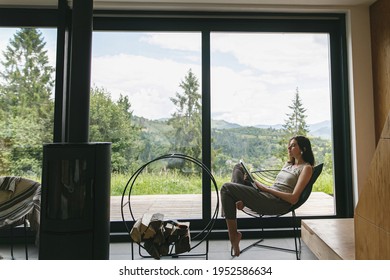 Beautiful stylish woman reading book on chair at fireplace with firewood on background of mountain hills. Young female in casual clothes relaxing in modern chalet with amazing view from window - Shutterstock ID 1952586634