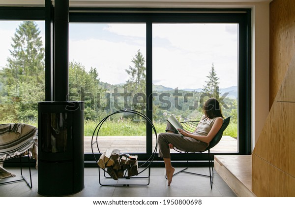 Beautiful stylish woman reading book on chair at\
fireplace with firewood on background of mountain hills. Young\
female in casual clothes relaxing in modern chalet with amazing\
view from window