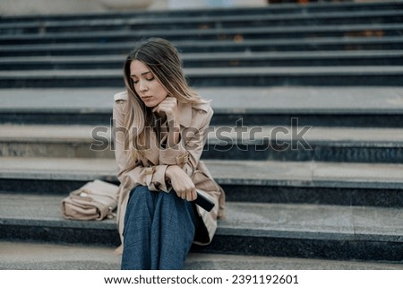 Beautiful stylish woman sitting on a stairs outside and looking really sad. Fashionable female in trench coat with a bag put on a side looking away and looking melancholic. Copy space.