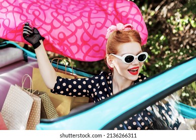 Beautiful Stylish Woman In Vintage Bright Dotted Clothes And Glasses Is Driving Mint Old Car After Shopping. Fashion Pin-up Girl Is Waving Pink Scarf. Retro Style Concept. Black Friday Sale.