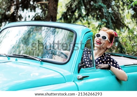 Beautiful stylish woman in vintage bright dotted clothes and glasses is driving mint old car after shopping. Fashion pin-up girl is travelling by auto. Retro style concept. Vacation trip mood.