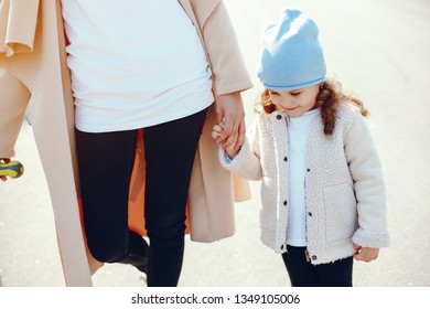 beautiful and stylish young mother with long dark hair dressed in a long coat and black pants walking in a sunny summer park with her little daughter in a white coat and hat. - Shutterstock ID 1349105006