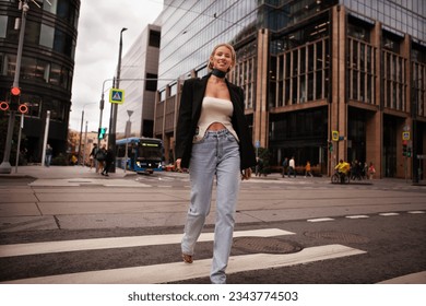 Beautiful and stylish young woman crossing road at pedestrian in city, smiling and dressed fashionably in jeans and blazer. Fashion street style on background of business center building.