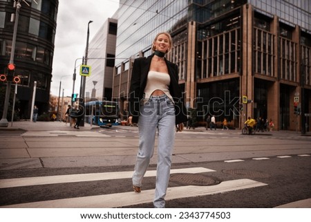 Beautiful and stylish young woman crossing road at pedestrian in city, smiling and dressed fashionably in jeans and blazer. Fashion street style on background of business center building.