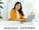 Beautiful successful brunette arabian or indian young woman in an orange shirt, IT specialist, product manager sits at a workplace working in a laptop on a project, looking at camera, smiles friendly