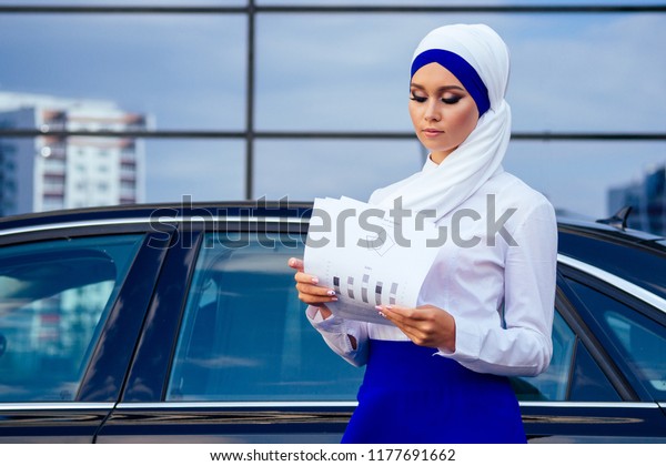 beautiful and successful european muslim business\
woman entrepreneur in stylish hijab and turban holding paper\
skyscraper office windows background on street standing near her\
black car