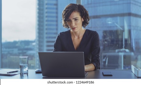 Beautiful Successful Female CEO Works On A Laptop In Her Modern Sunny Office With Cityscape Window View. Strong Female Business Leader.