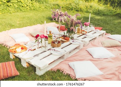 Beautiful summer celebration picnic in the garden with tasty food, cold drinks. Pallet table, Flowers decoration. Pleasure time with friends. Leisure and fun. White and pink colors