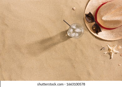 Beautiful Summer Holiday Beach Background With Straw Hat, Sunglasses, Glass Of Water And Shells On Sand Background, Top View With Copy Space. Harsh Shadows