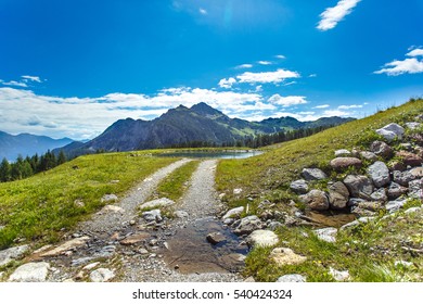 A beautiful summer landscape in the Alps with the mountain road. Fabulous meadow, mountain stream and lake, pine trees, large rocks and amazing clouds flying over on bright blue sky. Austria, Europe.
