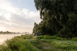 Beautiful Summer Landscape By The River In The Early Foggy Morning. The Tent Stands Among Green Grass And Trees, On The Shore Of A Lake. Camping. Watching Dawn Near A River Covered In Thick Fog