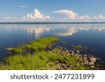 Beautiful summer landscape. View from the shore to the lake. Windless weather, calm. Clouds are reflected in the water. Rocky shore covered with grass. Lake Shotozero, Republic of Karelia, Russia.