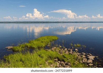 Beautiful summer landscape. View from the shore to the lake. Windless weather, calm. Clouds are reflected in the water. Rocky shore covered with grass. Lake Shotozero, Republic of Karelia, Russia. - Powered by Shutterstock