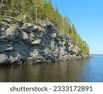 Beautiful summer nature of northern lake. Landscape with rocky coast, green trees, blue sky and reflection in calm water. Ladoga lake, Karelia, Russia.