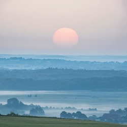 Beautiful Summer Sunrise Landscape Image Looking Over Fields With Mist In Distance And Hazy Sun Low In The Sky