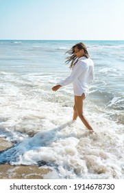 Beautiful Summer Woman Walking In Sea Water With Waves At Hot Sunny Day, Wearing Flowy White Shirt. Freedom And Paradise Concept