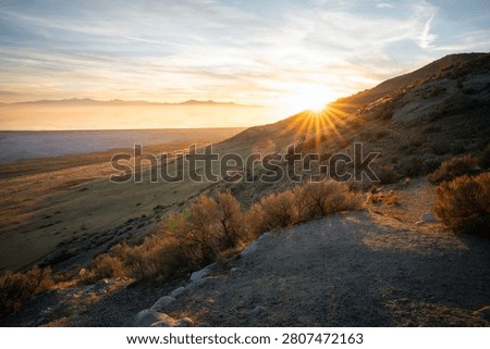Beautiful sunburst effect with sharp sun rays breaking over a rocky hill slope at Antelope Island, Utah. Warm golden hour light illuminating desert bushes and hiking trail at sunset.
