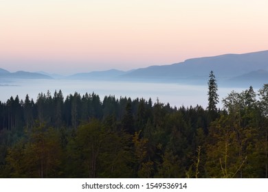 Beautiful Sunrise In The Mountains. Small Houses In A Meadow Among A Cone Of Coniferous Trees In The Fog. Great Awakening.