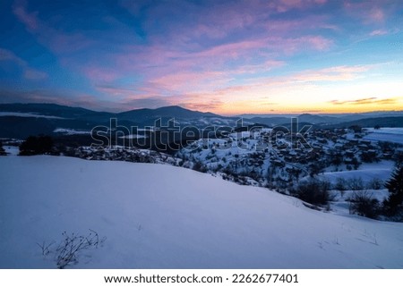 Beautiful sunrise view with snowy mountain slopes and small village among them in the frozen winter morning, the Rhodopi Mountains, Bulgaria 	