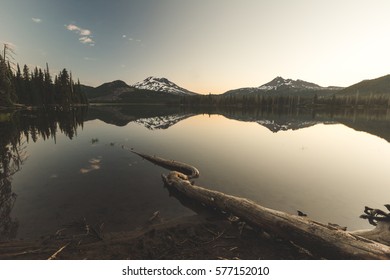 Beautiful Sunrise View Of Sparks Lake Near Bend, Oregon. Large Tree Root In Foreground With Two Mountain Peaks In Distance. Morning, Summer Or Spring Setting.