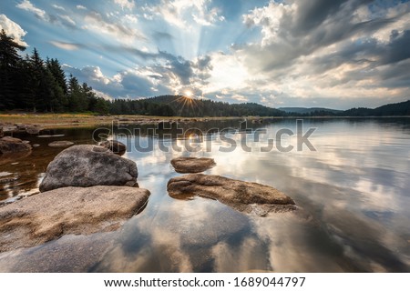Beautiful sunrise view with sun rays over a mountain lake surrounded by conifers. Shiroka Polyana dam in Rhodopi Mountains, Bulgaria.