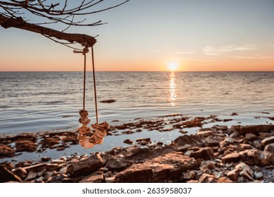 Beautiful sunset by the sea with hanging seashell decoration on driftwood branch and rocky beach. Warm light over tranquil seascape.