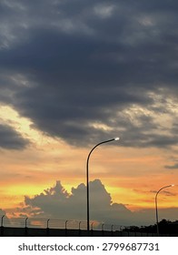 Beautiful sunset with dramatic orange and gray clouds over a road. Two curved streetlights are in the foreground, creating a striking contrast.