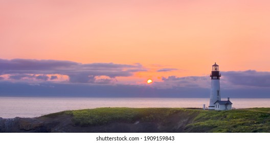 A beautiful sunset forms as the sun set below the horizon of the ocean with a lighthouse overlooking the grassy headlands of the coastline. 
