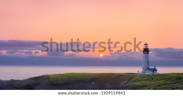 A beautiful sunset forms as the sun set\
below the horizon of the ocean with a lighthouse overlooking the\
grassy headlands of the coastline.\
\
