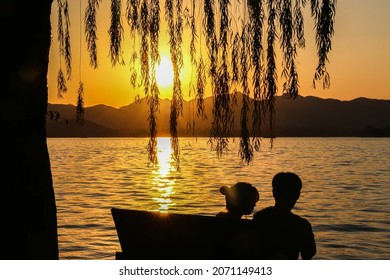 Beautiful sunset at Hangzhou West Lake, romantic silhouette of couple on bench enjoying the sunset by willow tree as narrow willow tree leaves fall from above