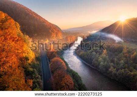 Beautiful sunset in mountains in autumn. Aerial view of forest with red trees on mountain, river, road, hills, village in fall in Ukraine. Colorful landscape. Top view from drone. Nature in october