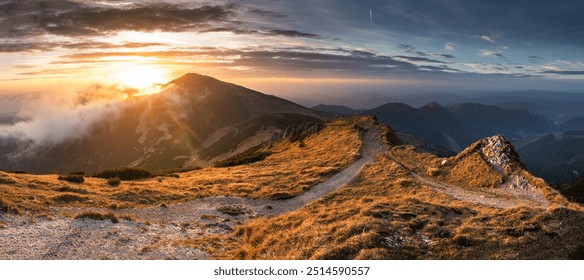 Beautiful sunset on mountain with trail leading up side. Trail is surrounded by grass and rocks. Sun is setting behind mountain, casting warm glow on landscape