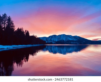 A beautiful sunset over the snowy mountains of Lake Tahoe reflects off the calm water. Many colors illuminate the sky behind the forest landscape