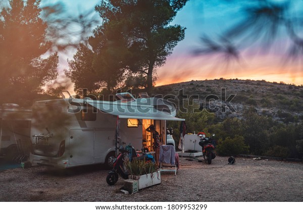 Beautiful sunset painted above\
bushy hill. Young woman is relaxing around campervan. She rode all\
day long her motorcycle and now she will rest in cosy\
campervan.