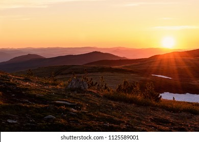 Beautiful sunset with rays over the contours of mountains with stones, small cedars and snow in the foreground - Powered by Shutterstock