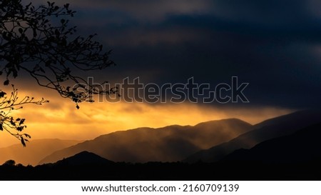 Beautiful sunset scene in Scottish Highlands. Dark clouds over silhouetted foggy hills lit by the golden setting sun and framed with a tree branch.