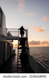 Beautiful Sunset Sky On Ferry Boat With Anonymous Passengers Climbing Steps Up Ladder To Top Deck Of Ship.  Sea And Horizon In View With Light Reflecting On Ocean.