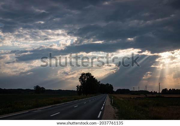 beautiful sunset with sun\
beams over the road. Sun behind dramatic clouds. Yellow\
agricultural field on road side. Road and white dividing lines make\
great perspective