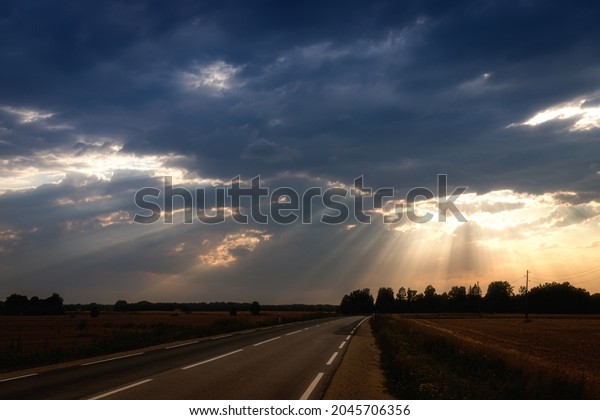 beautiful sunset with sun beams over the road. Sun\
behind dramatic clouds. Yellow agricultural field on road side.\
Road and white dividing lines make great perspective. Local\
electric poles and\
wires