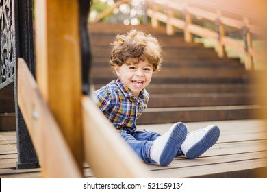 Beautiful Sunshine Toddler Boy Sitting On Old Wooden Stairs And Laughing Joyfully While Looking At Camera. Portrait Of Adorable Happy Baby Playing Alone Outside On Warm Summer Day. Horizontal Image.